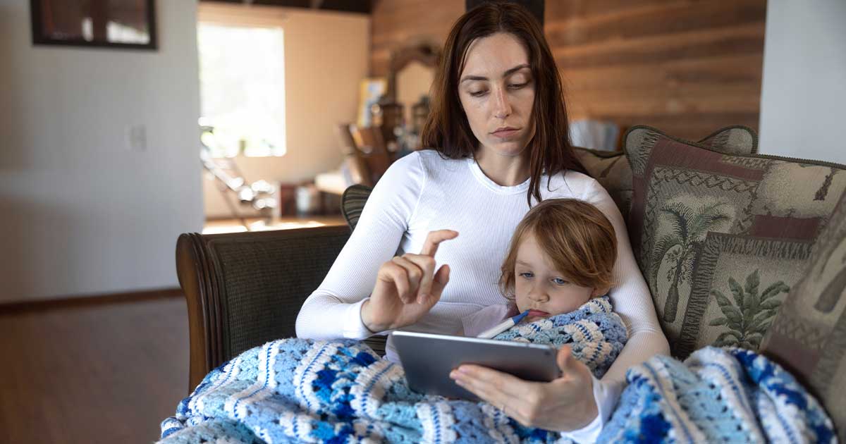 Frau mit krankem Kind auf Couch hält Tablet und tippt.