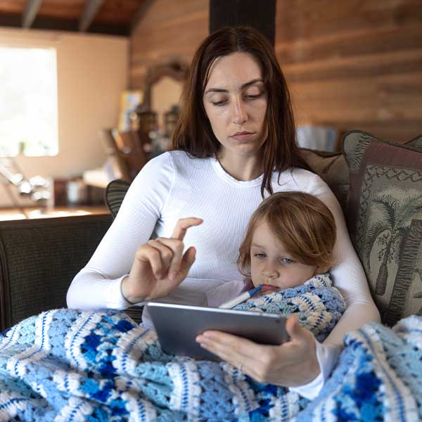 Frau mit krankem Kind auf Couch hält Tablet und tippt.