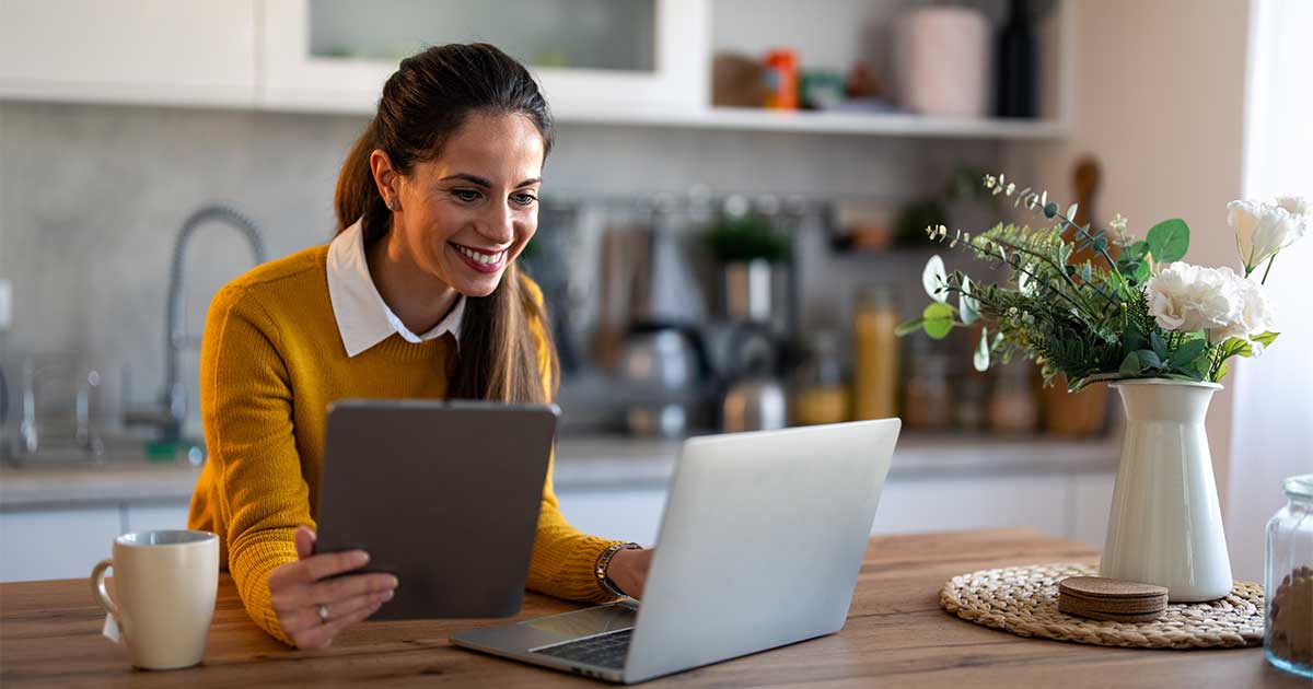 Frau lehnt an Tisch, hält ein Tablet in der Hand und schaut auf einen Laptop.